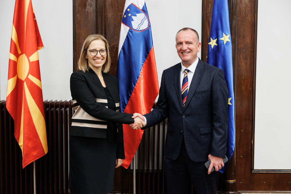 Ministrica in minister se rokujeta pred slovensko, makedonsko in zastavo Evropske unije. Ministrica ima črn jopič z belimi črtami in črno krilo, minister pa temno modro obleko z modro-rdečo-rumeno kravato.