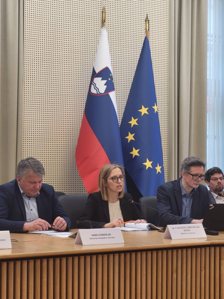 Ministrica sedi za leseno mizo, ob njej sedijo še trije moški. Ministrica govori v mikrofon. Za njo sta slovenska zastava in zastava Evropske unije.