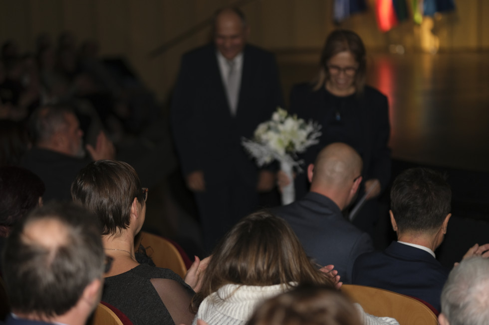 Ministrica s šopkom rož hodi proti praznemu stolu v dvorani. Vsi drugi stoli so že zasedeni in ljudje ploskajo.