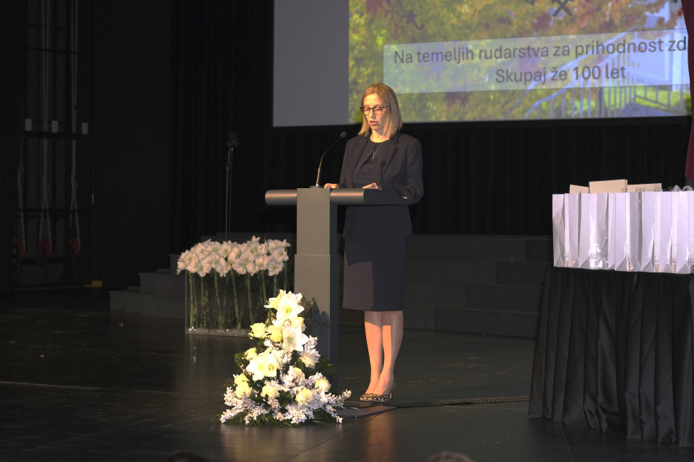 Ministrica stoji za govorniškim pultom. V ozadju se vidi del projekcije, pred govornico na tleh je bela ikebana.  