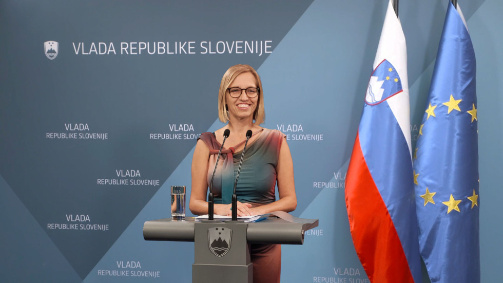 Ministrica za zdravje v pisani poletni obleki z nasmeškom stoji za govornico, v ozadju stena z napisom Vlada RD in slovenska zastava