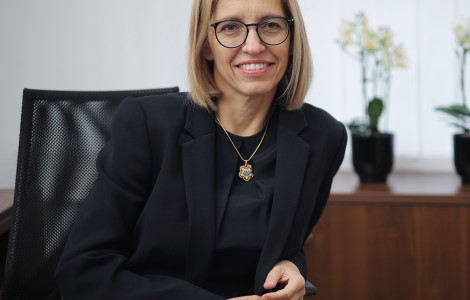 Ministrica (The minister is sitting at a table and laughing. There are papers and a pencil on the table. Behind her is a cabinet with white orchids on it. The minister is wearing a black T-shirt and a black jacket. She is wearing a gold chain. She has glasses.)