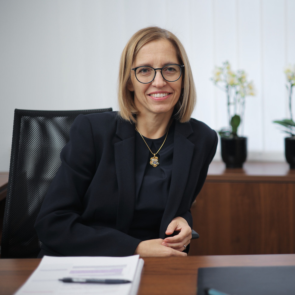 The minister is sitting at a table and laughing. There are papers and a pencil on the table. Behind her is a cabinet with white orchids on it. The minister is wearing a black T-shirt and a black jacket. She is wearing a gold chain. She has glasses.