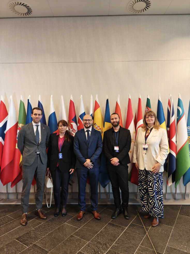 Skupina odraslih oseb v poslovnih oblačilih se slika pred zastavami