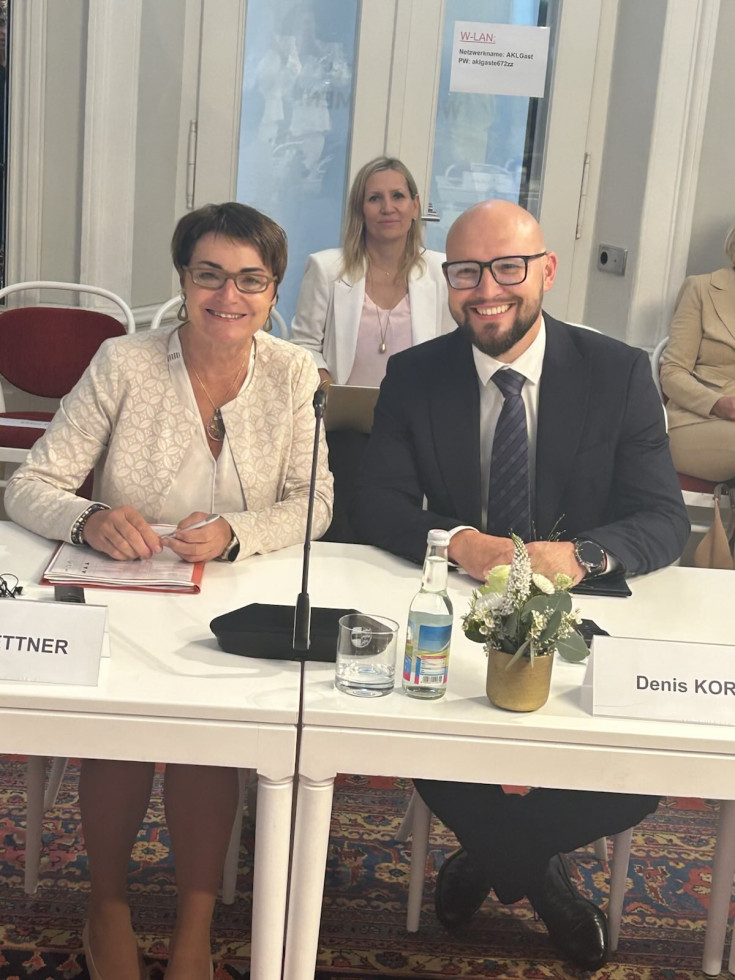Moški v temni poslovni obleki z očali, sedi za mizo z žensko v bež poslovni obleki z očali in kratkimi lasmi. V ozadju plavolaska ženska v poslovni obleki