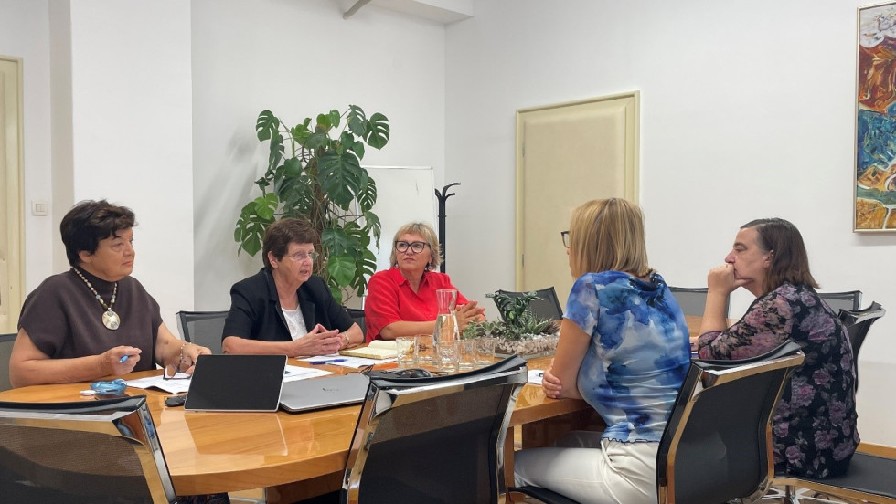 Ministrica sedi v svetlo modri majici in belih hlačah, ob njej ženska v cvetlični majici, nasproti za mizo sedijo še 3 starejše gospe. 