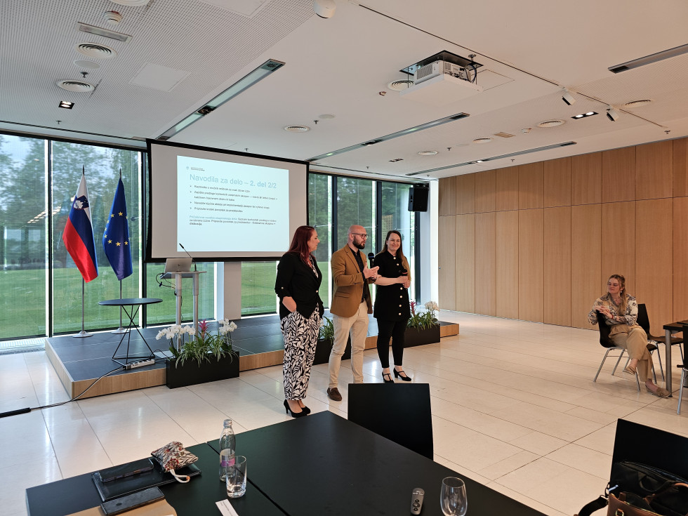 V dvorani stojijo dve ženski in moški. Moški govori v mikrofon. Za njimi je nizek oder, na njem govorniški pult, miza, slovenska zastava in zastava Evropske unije. Pred odrom so tri korita z rožami. Stena zadaj je steklena, zunaj pa trava in drevesa.