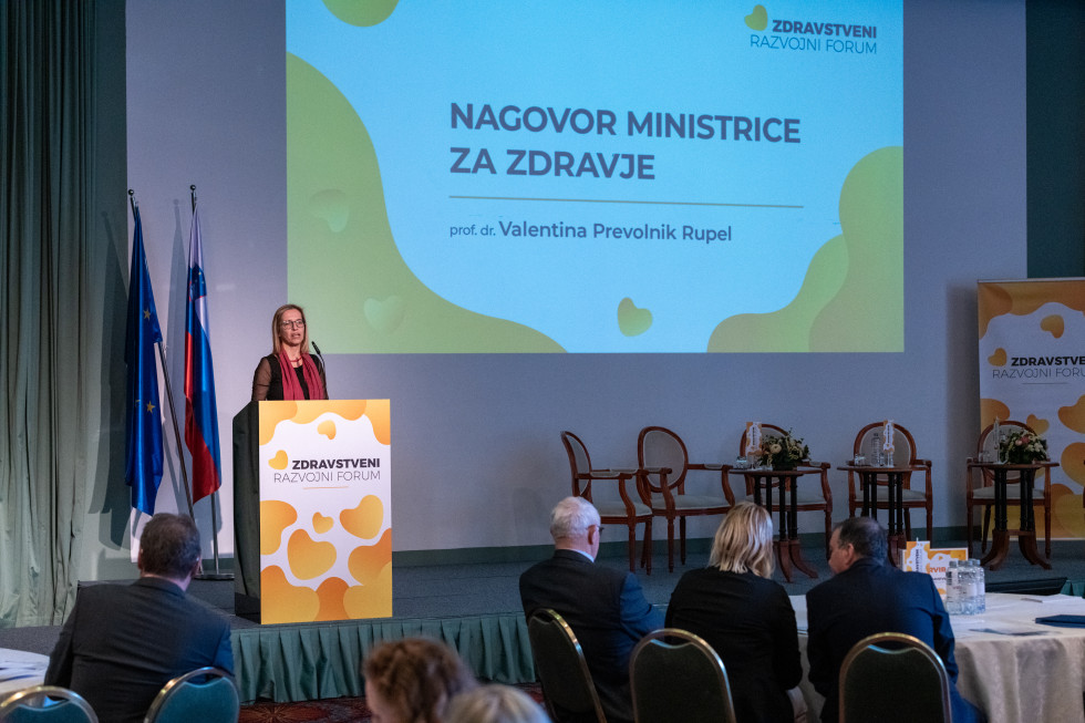 Ministrica stoji na odru za govorniškim pultom, za njo sta slovenska zastava in zastava Evropske unije, na steni pa projekcijsko platno z njenim imenom. Na odru so še prazni stoli in mizice. Pred odrom ljudje sedijo za okroglimi mizami in poslušajo.