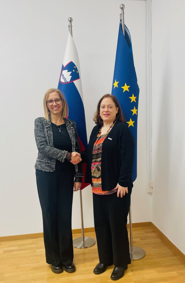 Ministrica in predstavnica urada se rokujeta pred slovensko zastavo in zastavo Evropske unije.