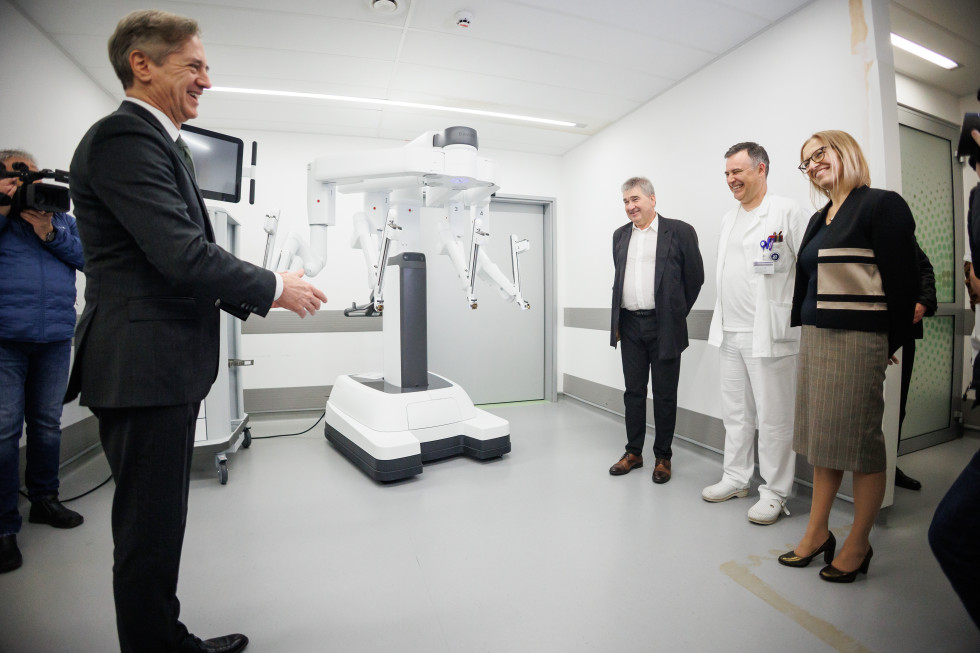 Ministrica si v beli bolnišnični sobi ogleduje nenavadno napravo. Ob njej stojita dva moška, eden od njiju je v beli zdravniški opravi.