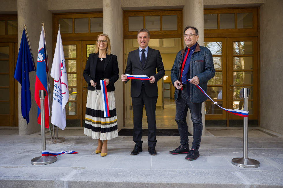 Ministrica, predsednik vlade in generalni direktor pred vhodom v bolnišnico simbolno režejo trak. Za njimi so velika stekleno-lesena vrata, ob njih pa tri zastave.