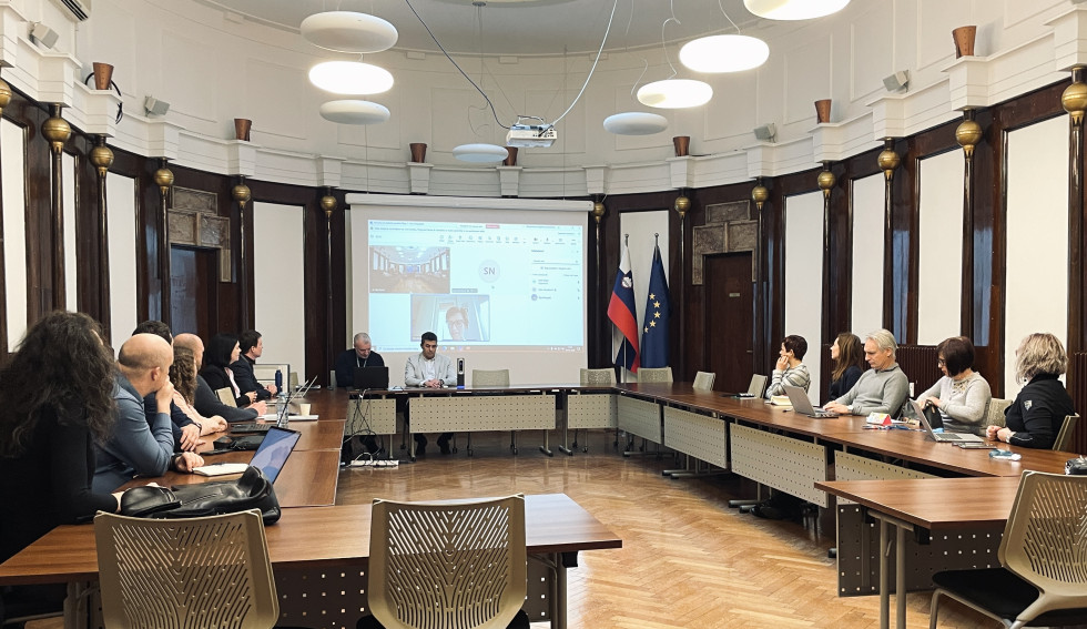 V veliki sejni sobi za mizami, sestavljenimi v črko U, sedijo ljudje in sestankujejo. Na steni je projekcijsko platno, v kotu pa slovenska zastava in zastava Evropske unije.