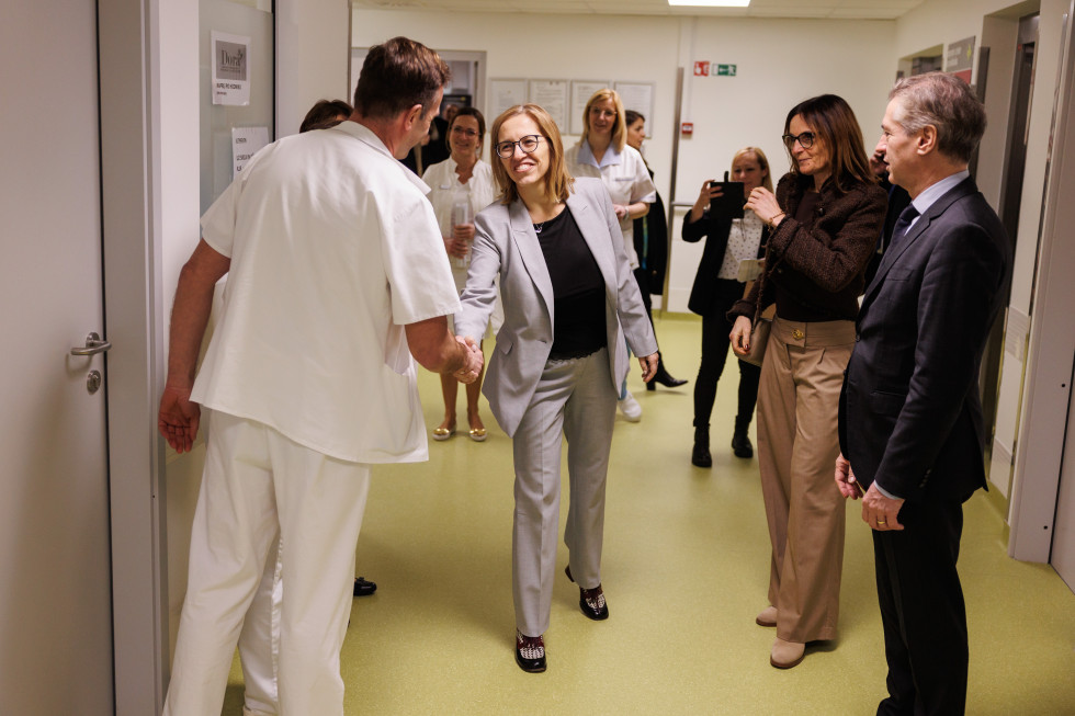 Ministrica se na bolnišničnem hodniku rokuje z moškim v beli zdravniški uniformi, zraven stoji premier, za njima pa še drugi ljudje, nekateri v zdravniških uniformah.