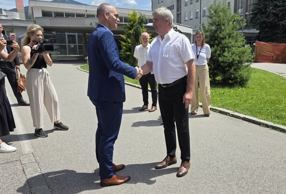 Pred vhodom bolnišnice se rokujeta minister in direktor. Okoli njiju stoji nekaj ljudi, med njimi tudi snemalka s kamero. 