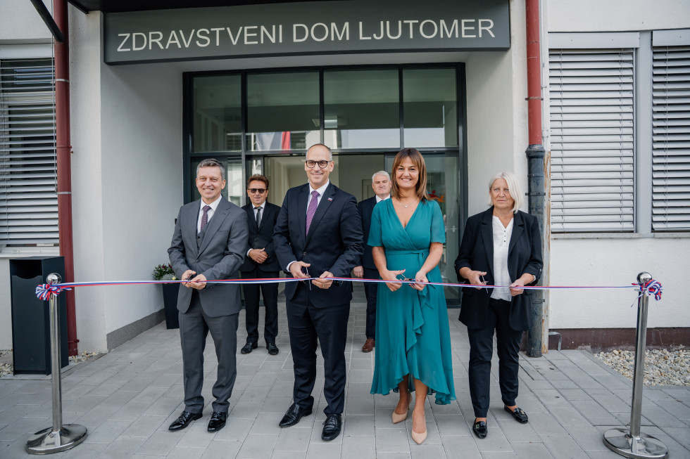 Pred vhodom v zdravstveni dom stojijo dva moška in dve ženski. V rokah imajo škarje in se pripravljajo na rezanje traku v barvah slovenske zastave. Za njimi stojita še dva moška.