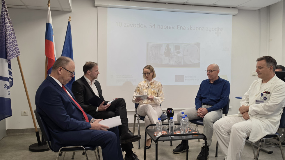 Štirje udeleženci okrogle mize sedijo zbrani pred občinstvo, moderatorka na sredini povezuje pogovor. 