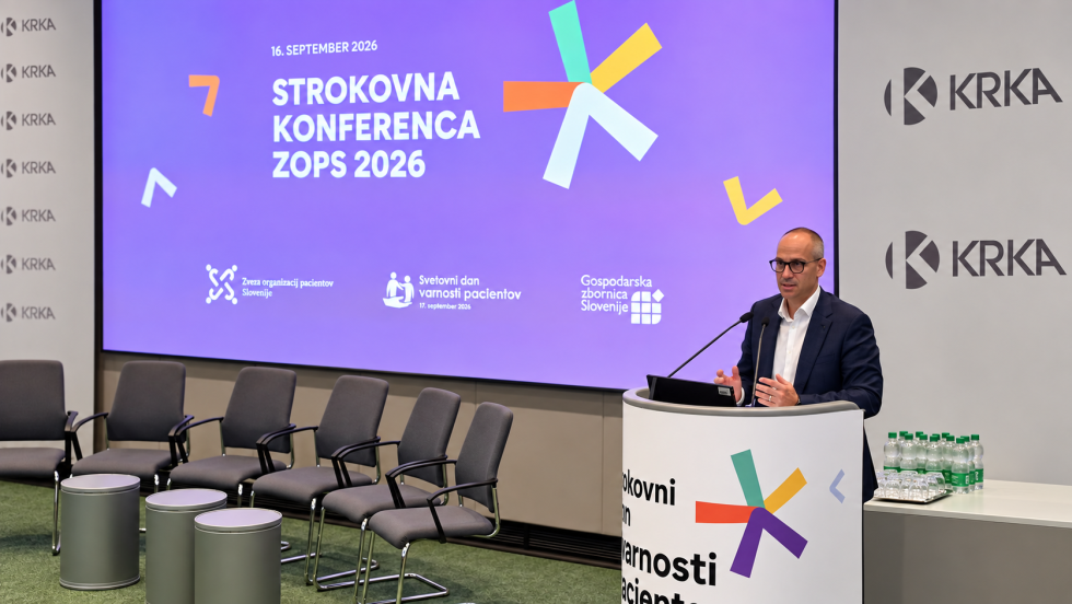 Minister za zdravje stoji na odru za govorniškim pultom in ima govor udeležencem. Za njim je projekcija z nazivom dogodka in logotipi organizatorjev