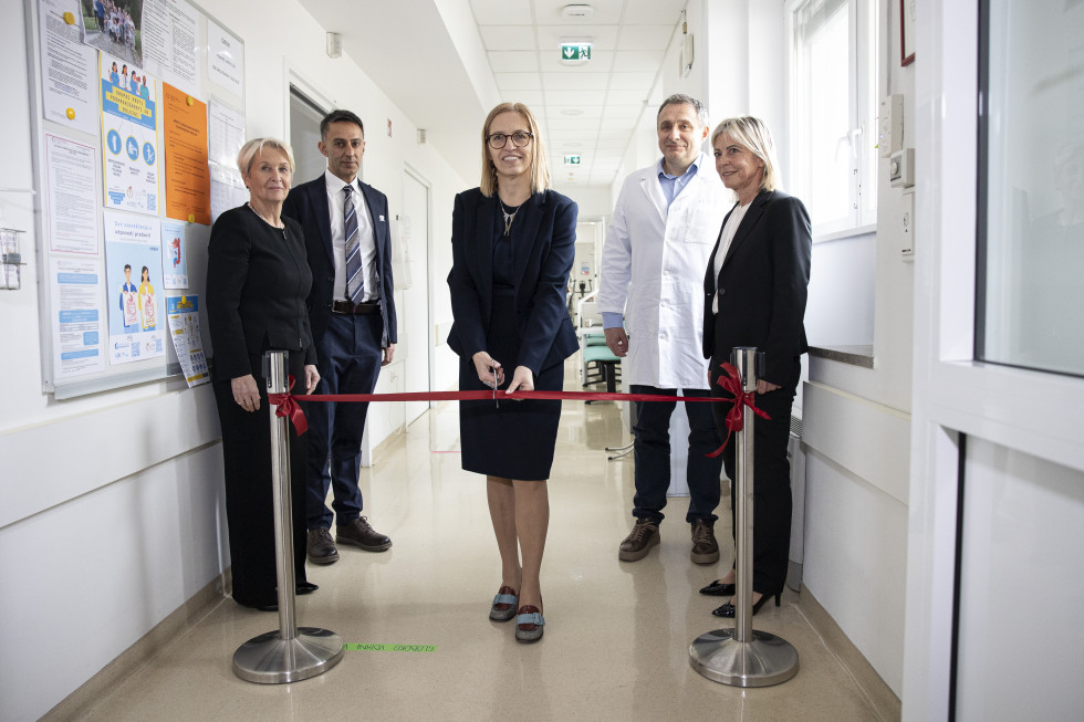 Ministrica na hodniku s škarjami reže rdeči trak, za njo stojita še dve ženski in dva moška, eden v beli zdravniški uniformi.