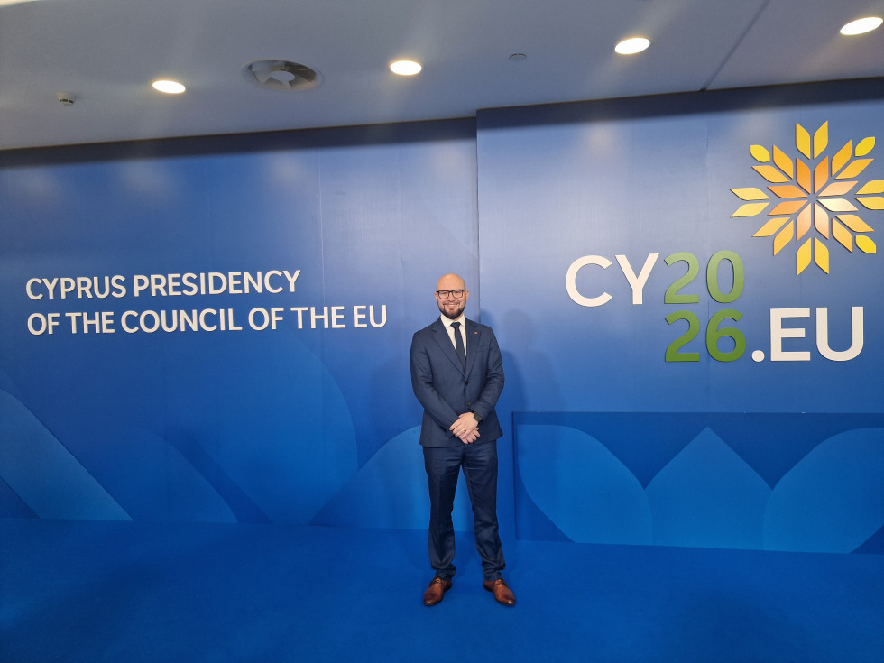 Državni sekretar pozira na modrem hodniku, za njim je logotip srečanja. Oblečen je v modro obleko, belo srajco in rjave čevlje, nosi kravato.