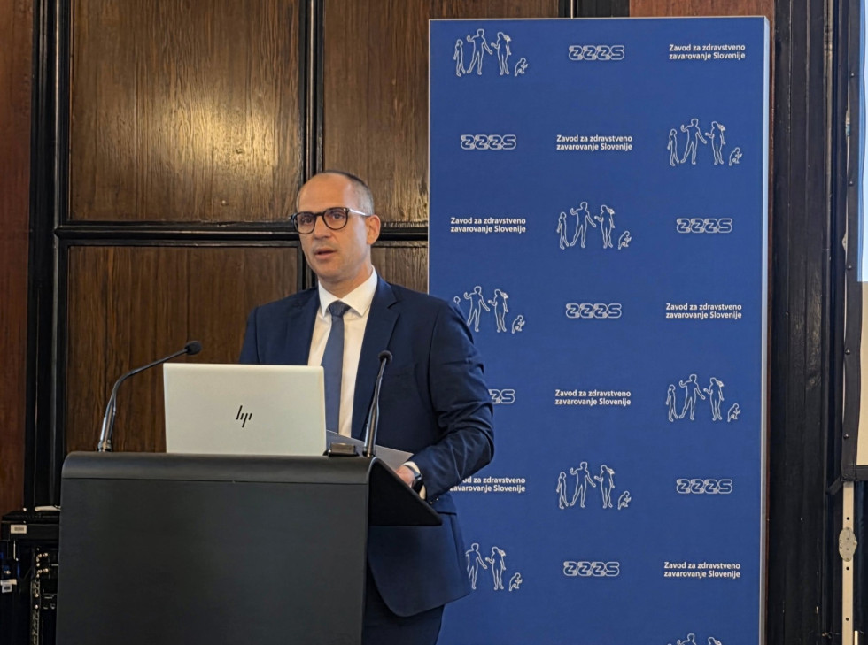 Minister za zdravje v temno modri obleki stoji za govorniškim pultom, za katerem stoji pano z napisi ZZZS, in nagovarja člane skupščine