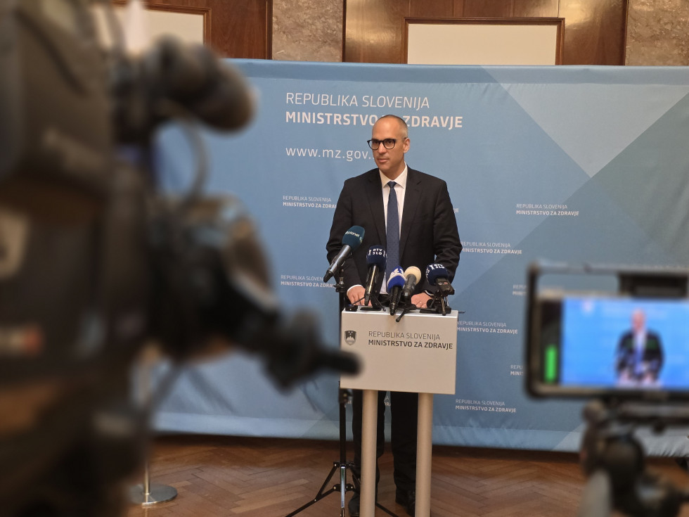 Minister stoji za govorniškim pultom, na katerem je več mikrofonov. Pred njim se zamegljeno vidijo kamere. Za njim je pano z logotipom Ministrstva za zdravje.