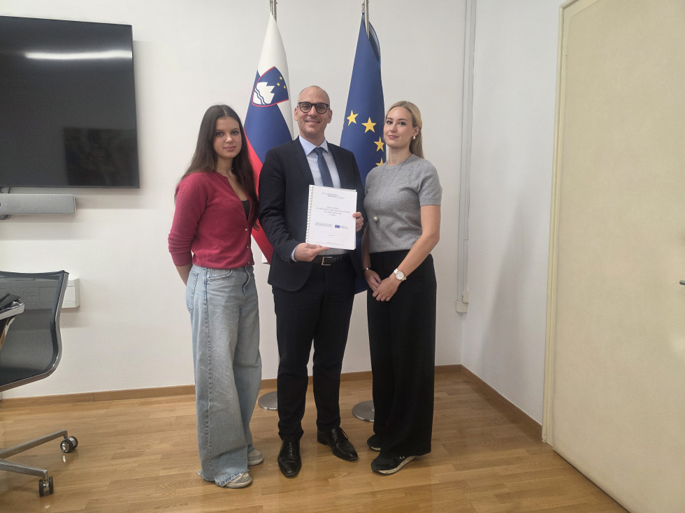 Minister stoji med dvema dekletoma, v rokah drži knjigo. Za njimi sta slovenska zastava in zastava Evropske unije.