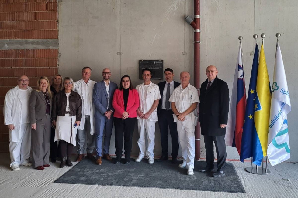 Skupina ljudi pozira pred betonsko-opečnatim zidom, nekateri so v belih zdravniških haljah. 