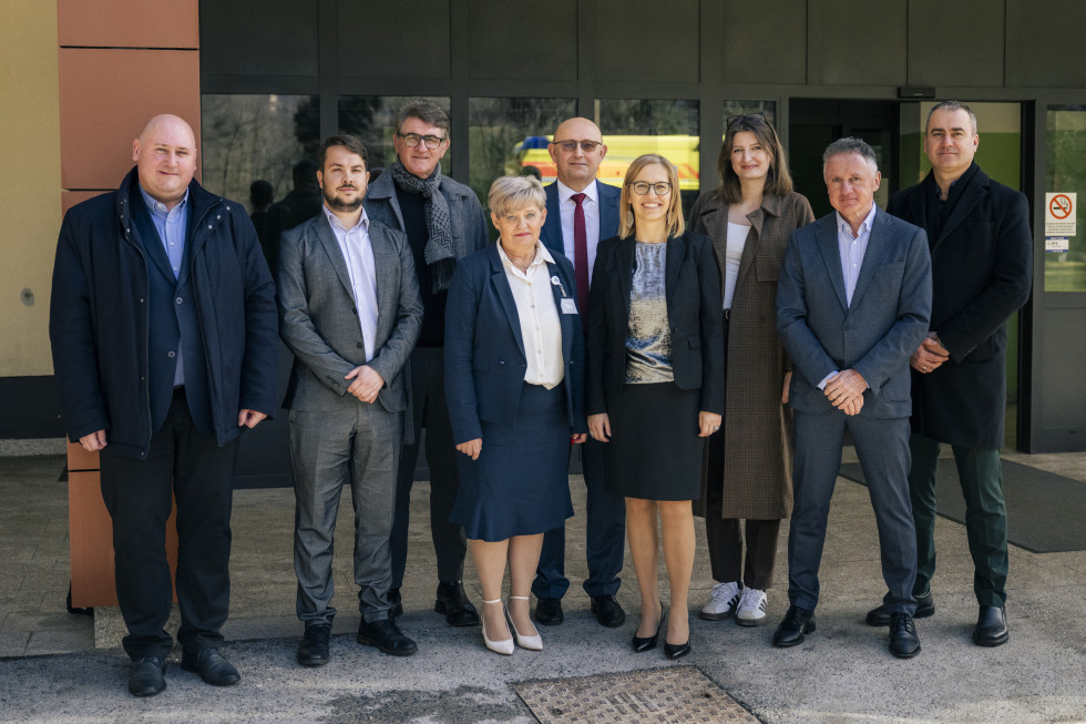 Ministrica pred vhodom v bolnišnico pozira z večjo skupino moških in žensk.