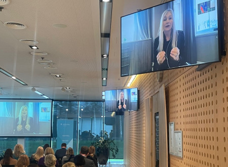 Na zaslonih dvorane je slika gospe z blond dolgimi lasmi, ki govori