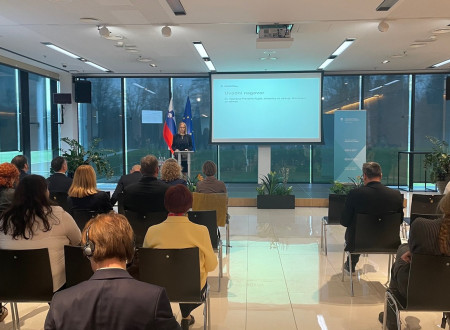 Ministrica stoji za govorniškim pultom, v ozadju zastave SLO in EU ter prezentacijski diapozitiv