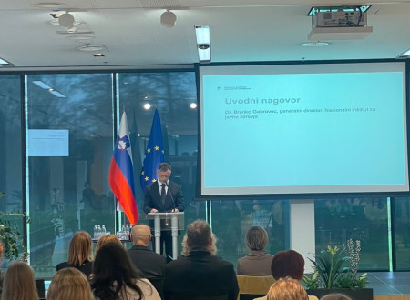 Moški v temni obleki stoji in nagovarja publiko v dvorani