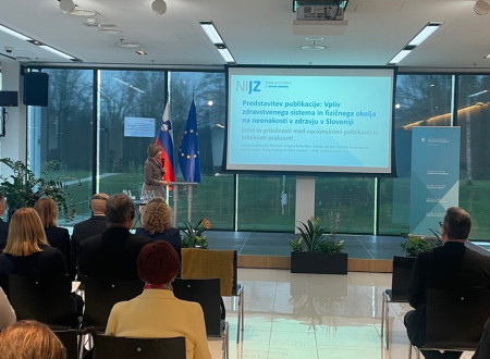 Gospa z mikrofonom v roki govori sedeči publiki z odra