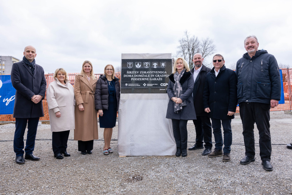 Zunaj na pesku ob temeljnem kamnu pozira več žensk in moških. Za njimi je oranžna varnostna gradbiščna ograja.