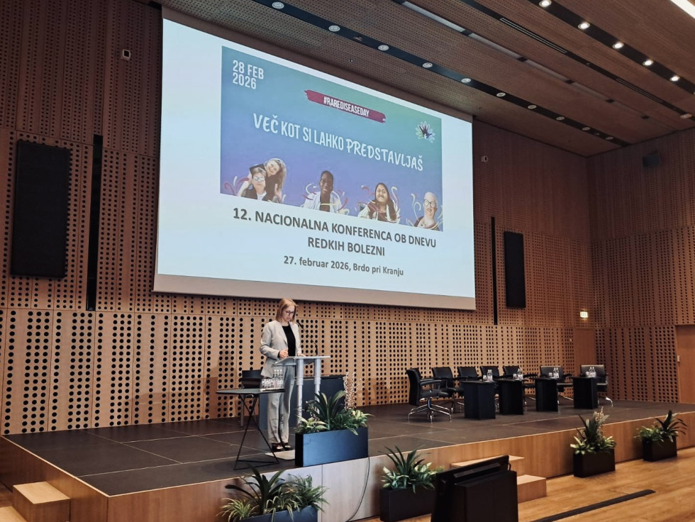 Ministrica stoji na odru za govorniškim pultom, ob strani odra so štiri mizice in več stolov. Ob odru so korita z rožami. Na steni je projekcijsko platno.