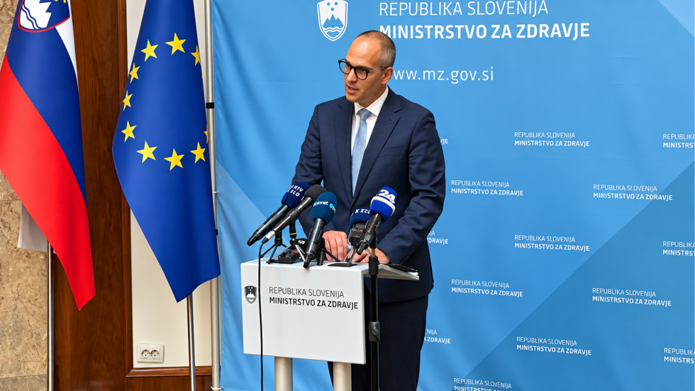 Minister stoji za govorniškim pultom, na katerem je več mikrofonov. Poleg njega sta slovenska zastava in zastava Evropske unije, za njim pa modra stena z napisom Ministrstvo za zdravje.