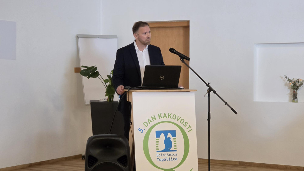 Za govorniškim pultom stoji moški. Govori v mikrofon.