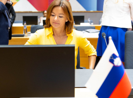 Ministrica Fajon sedi v dvorani za računalniškim zaslonom in s slovensko zastavo. 
