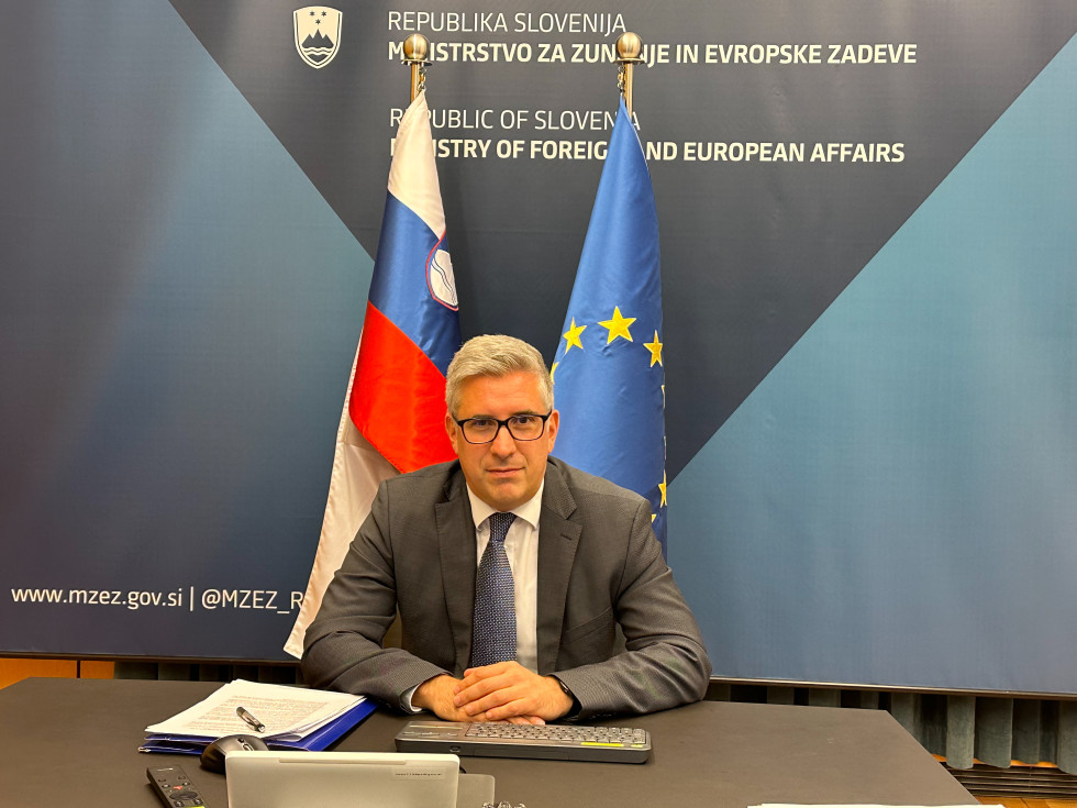 državni sekretar sedi pred slovensko in EU zastavo in gleda naprej
