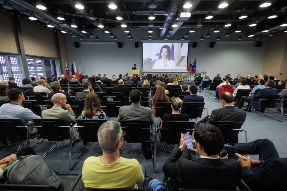 udeleženci konference, slikani iz ozadja dvorane, spredaj velik zaslon, na katerem se predvaja nagovor ministrice Fajon