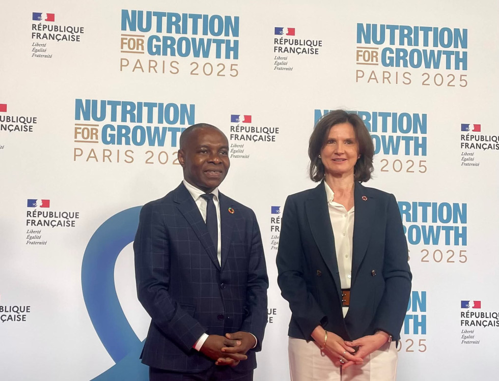 Moški in ženska stojita eden zraven drugega. Zadaj reklamni pano z napisom Nutrition for growth Paris 2025