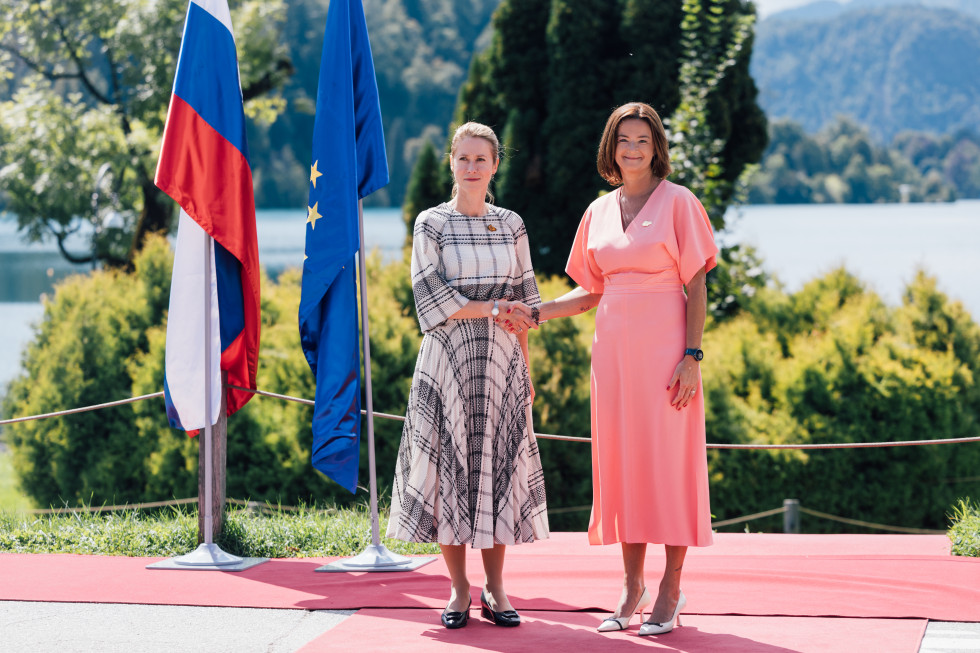 obe stojita na rdeči preprogi, v ozadju blejski otoček, na levo slovenska in EU zastava