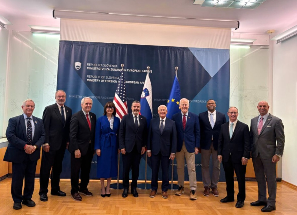 Ten people dressed in formal business attire are posing in a large office in front of the flags of the United States, Slovenia, and the European Union