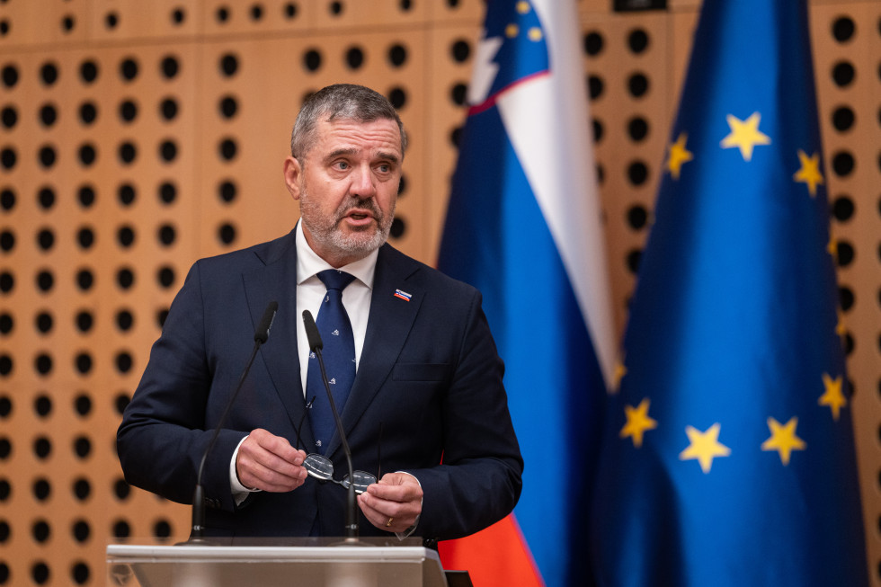 Minister med nagovorom na posvetu, za govorniškim pultom, zadaj slovenska in evropska zastava.