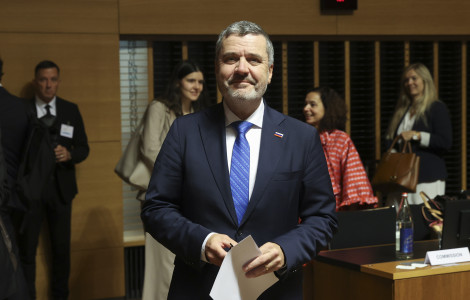 e493e9b0 dbae 4fbf 9cac 23610c686850 (A suited man holding papers stands in a conference room, with colleagues and a “Commission” nameplate behind him.)