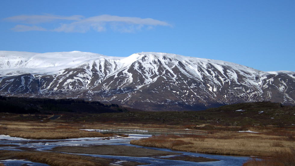 a small river flowing in brown grass covered land, snow covered mountains in the back