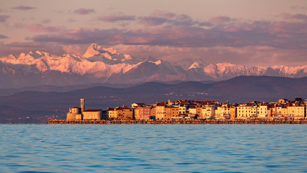 coastal city of Piran at sunset; sea at the forefront, snow covered mountains in the background