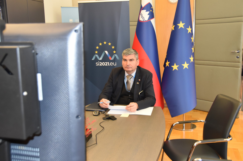 državni sekretar Raščan sedi za mizo, v ozadju zastave, AVK pogovor