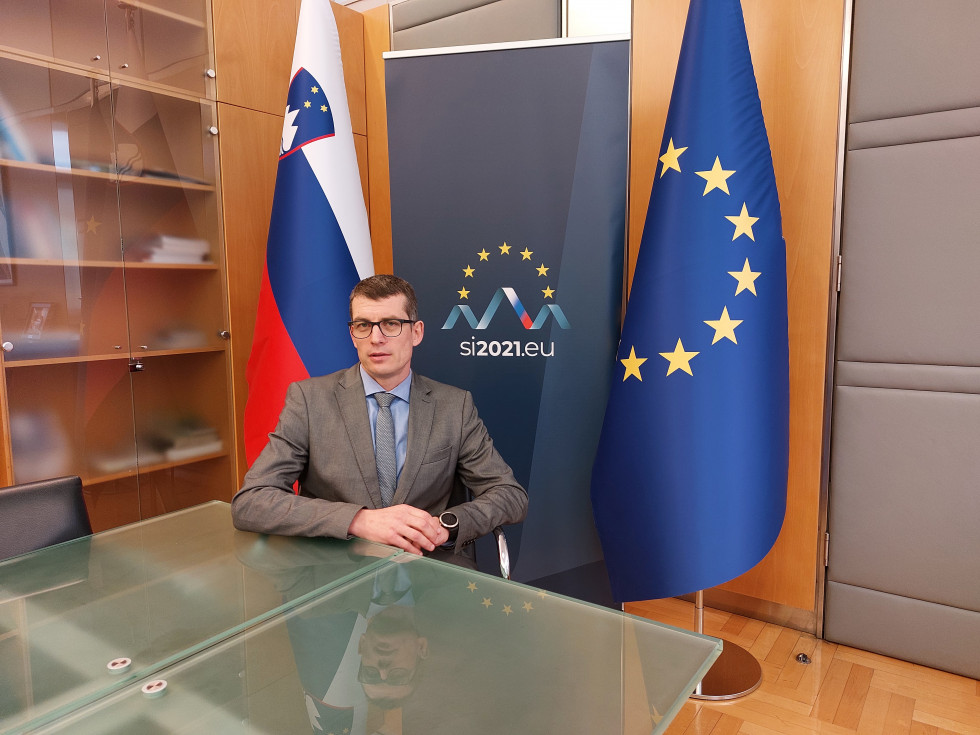 državni sekretar Dovžan sedi za mizo, za njim EU in slovenska zastava