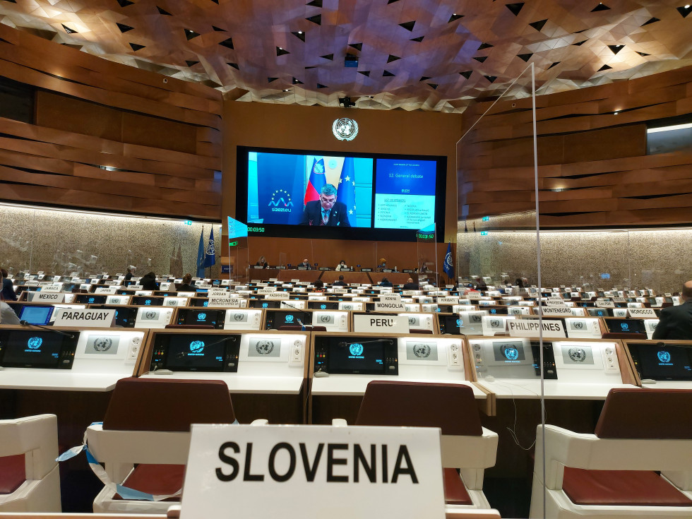 državni sekretar med zasedanjem, sedi na stolu, v ozadju slovenska zastava in pano z logom slovenskega predsedstva