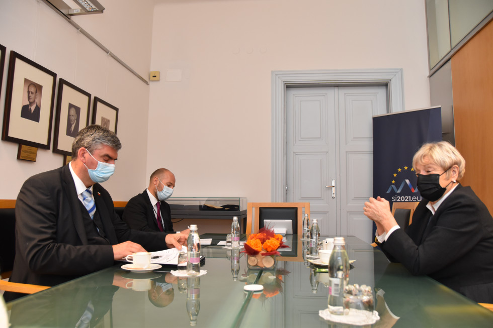 državni sekretar sedi za mizo, pogovor s sogovornico, v ozadju pano slovenskega predsedovanja